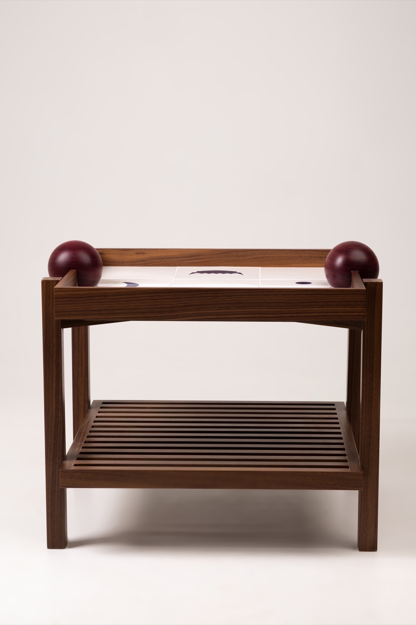 Mesa Lateral Sobral vista de frente: estrutura de madeira maciça sobre quatro pés, prateleira inferior ripada e tampo revestido de azulejo branco, com uma esfera de Roxinho encaixada em cada extremidade do tampo.